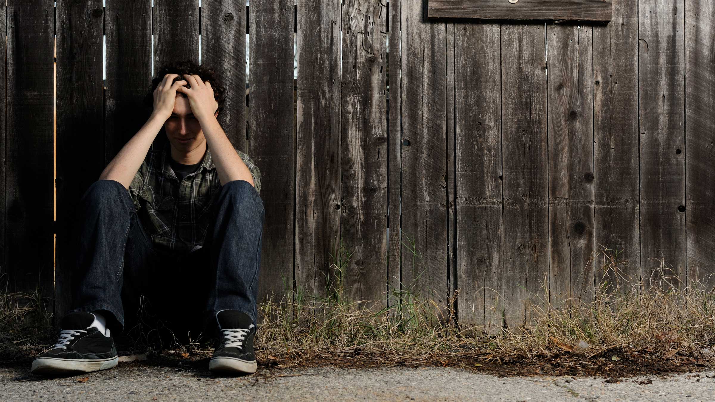 Lonely teenager sits against a fence with his head in his hands