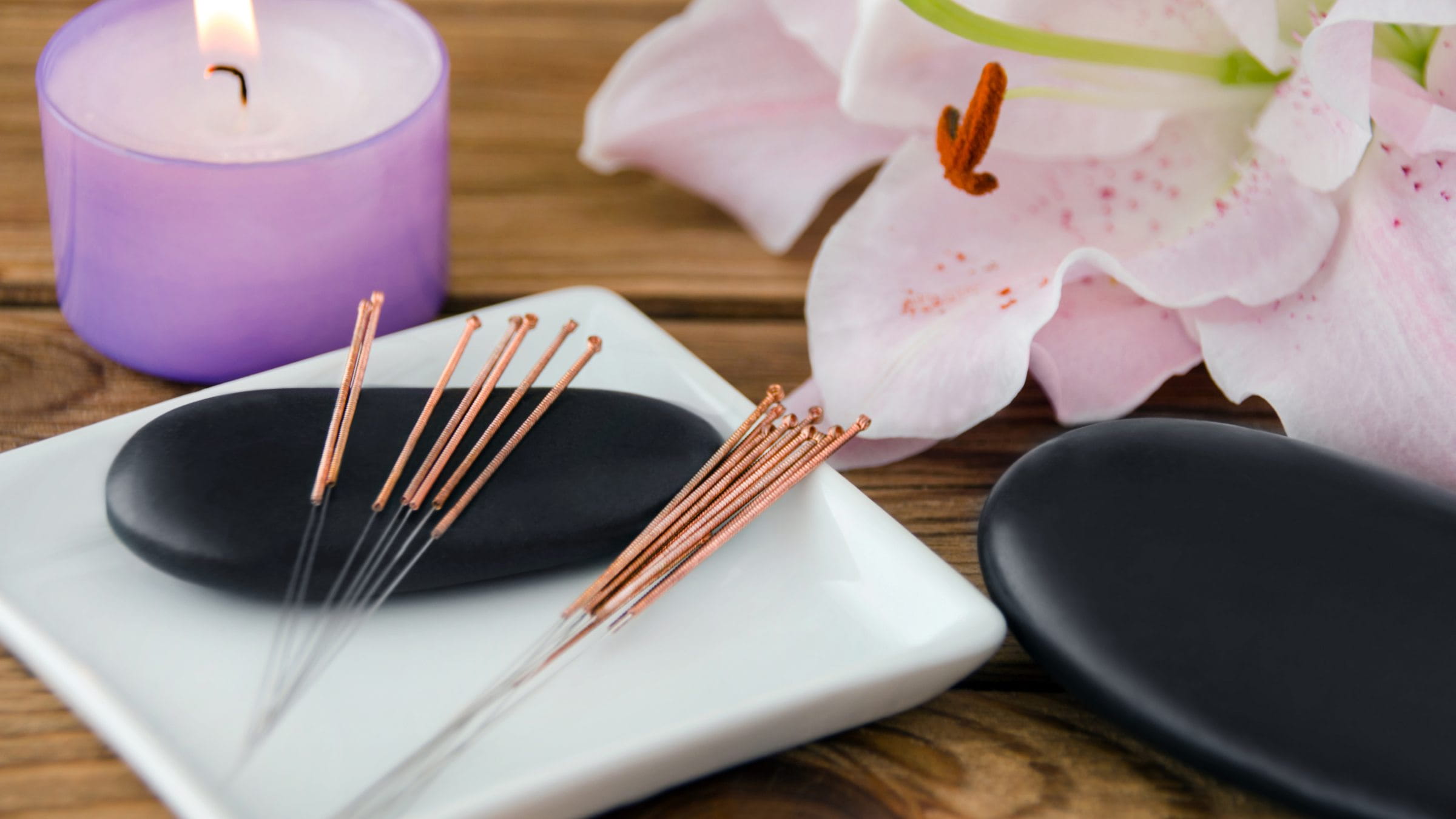 Close up image of a burning candle, acupuncture needles, stones and a flower resting on a wooden surface.