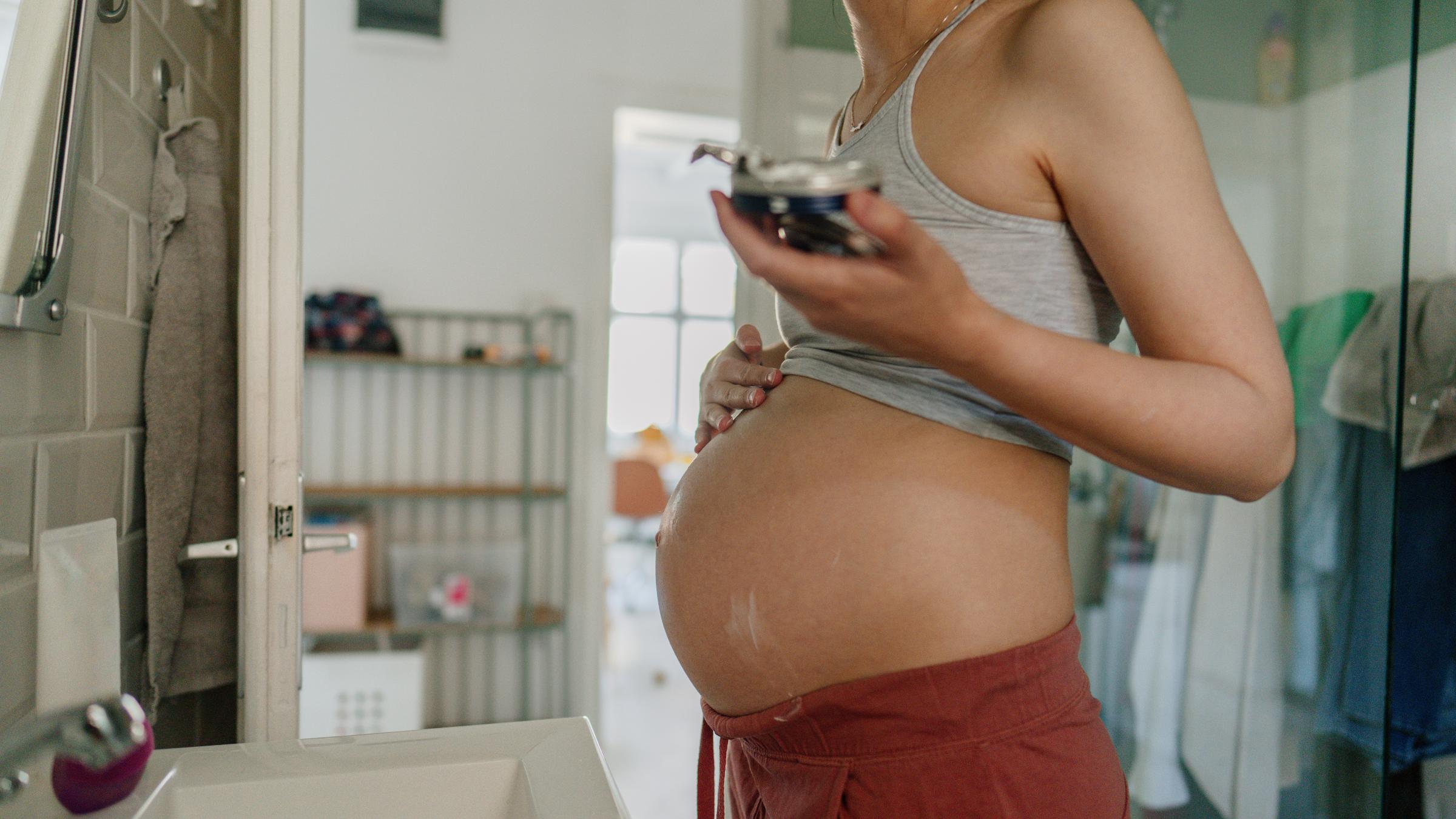 A pregnant woman standing in a bathroom rubbing topical cream onto her stomach