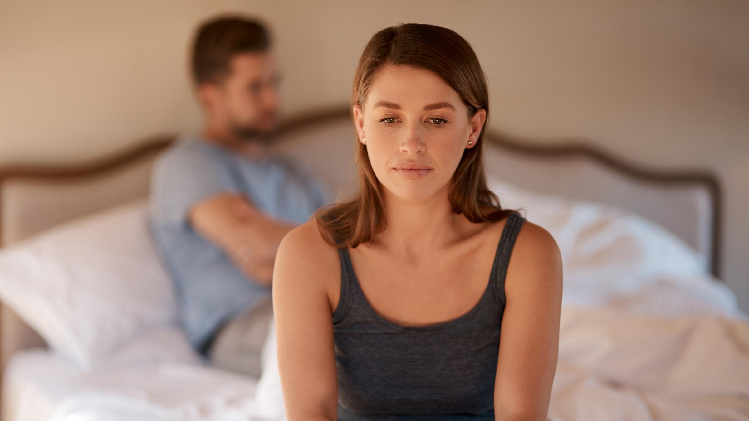 Woman setting on the edge of the bed with a man on the background