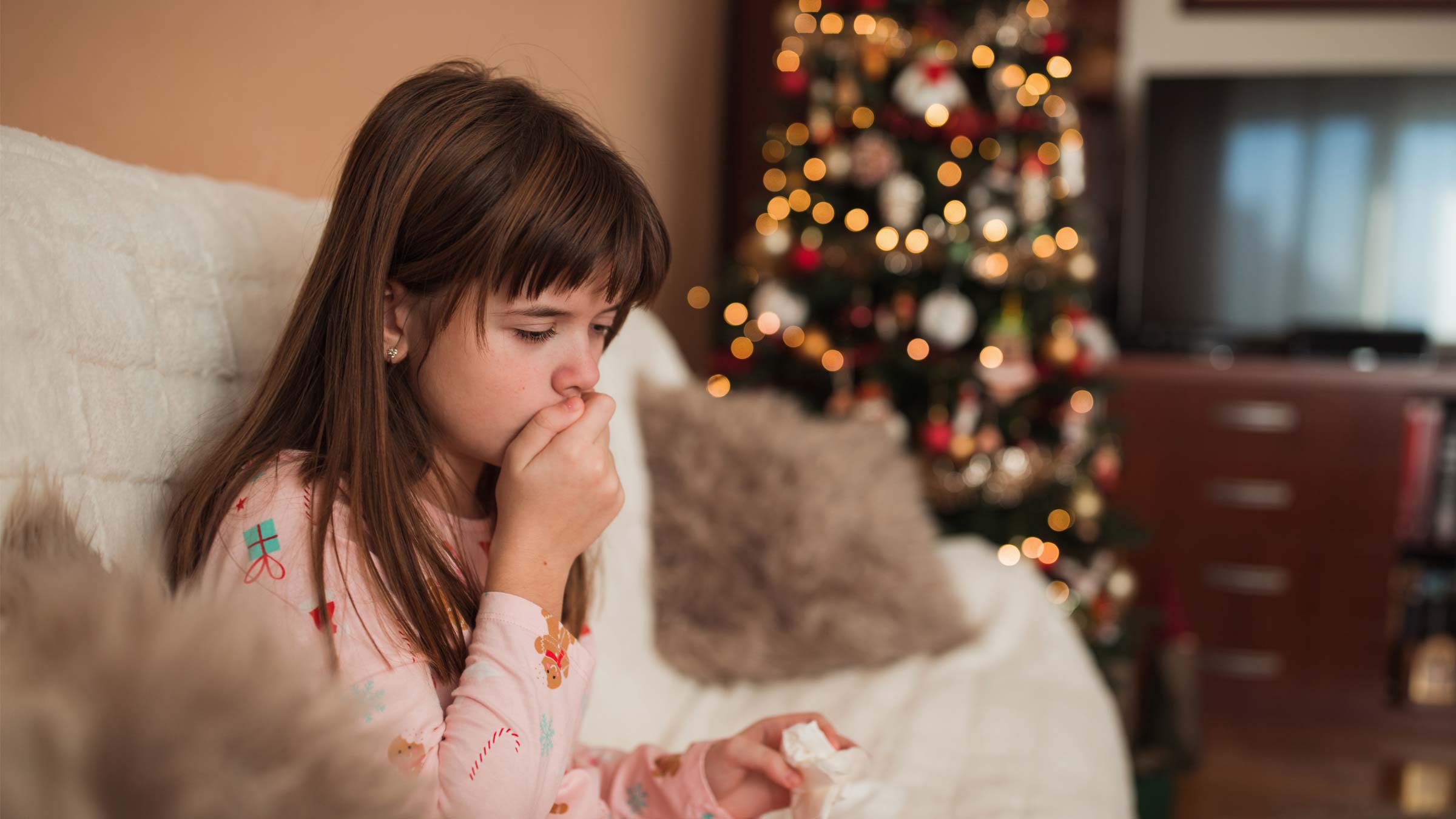 Girl sitting near Christmas tree holding hand over mouth when coughing