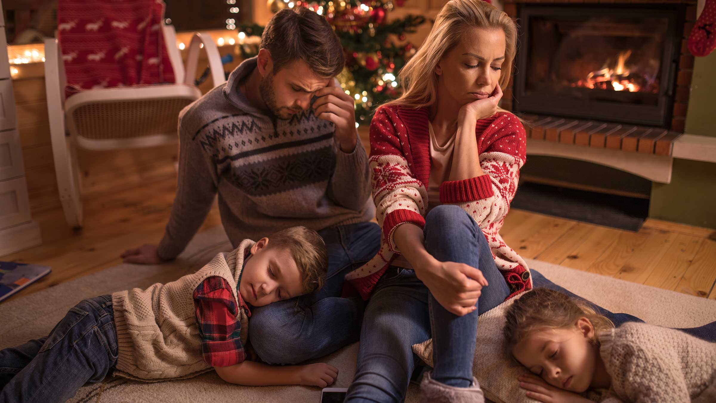 Parents appear annoyed at each other while two kids sleep at their feet in living room with holiday tree. 