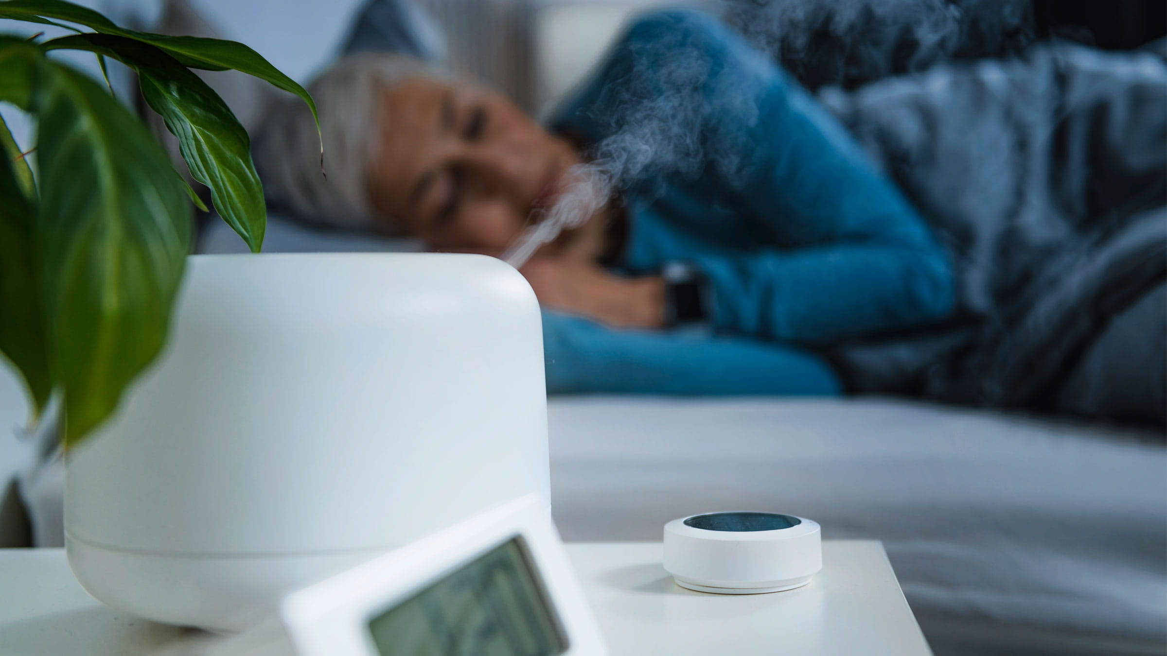 A woman appears to be sleeping in bed while a humidifier emits moisture into the air in the foreground.