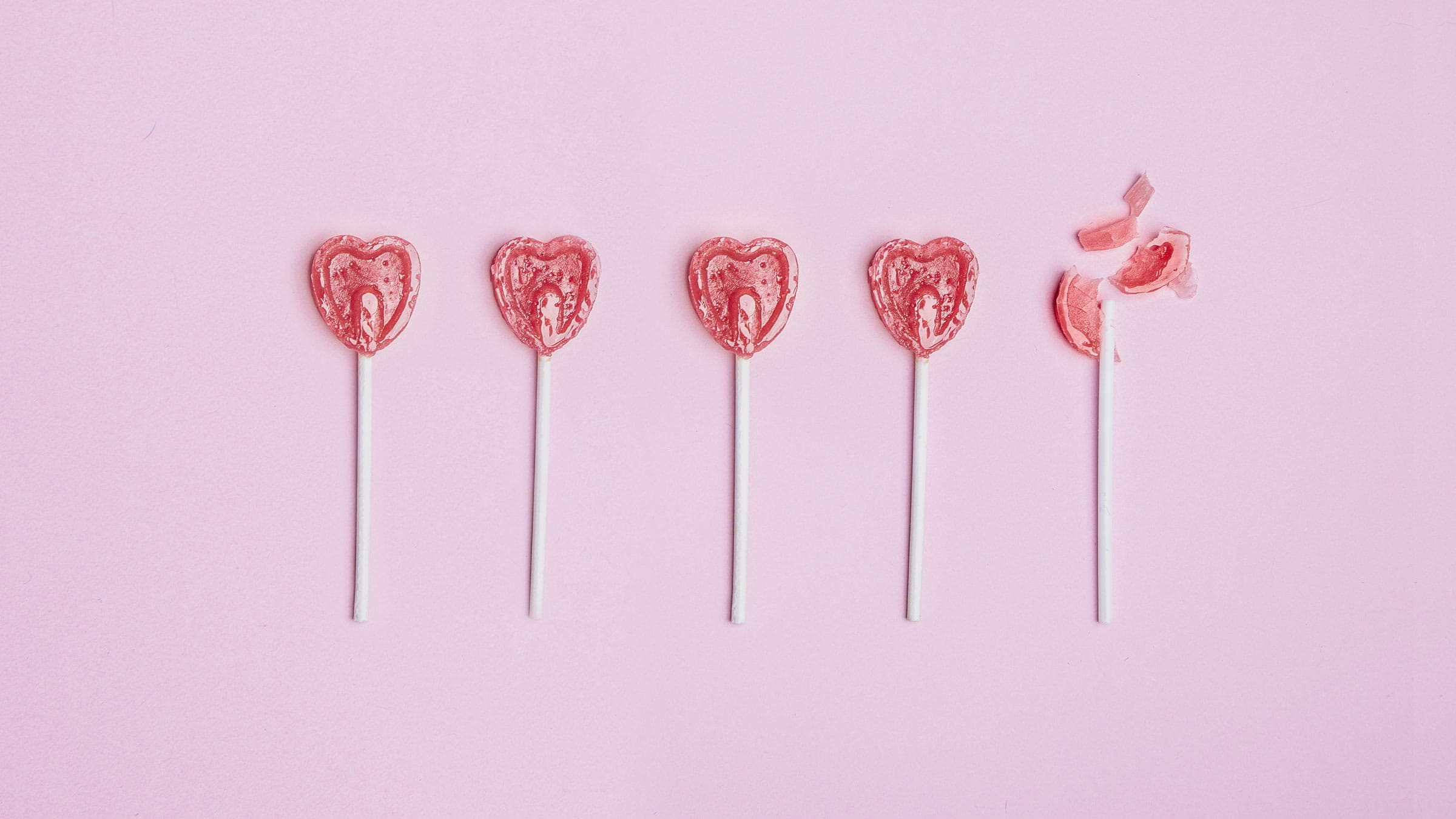 Five red, heart-shaped lollipops on a pink background. The last lollipop is cracked.