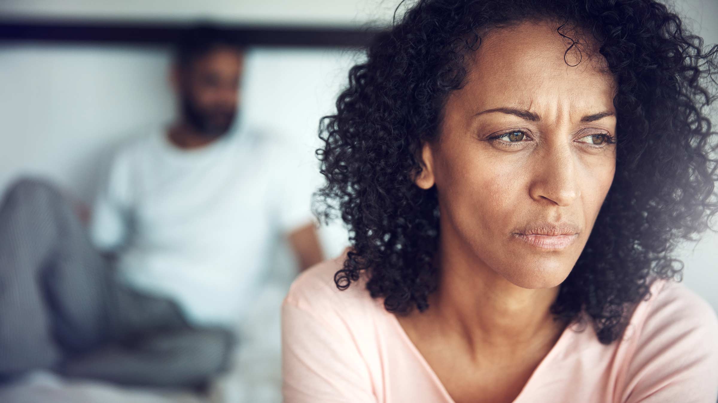 Concerned woman sitting on the edge of the bed with man in the background