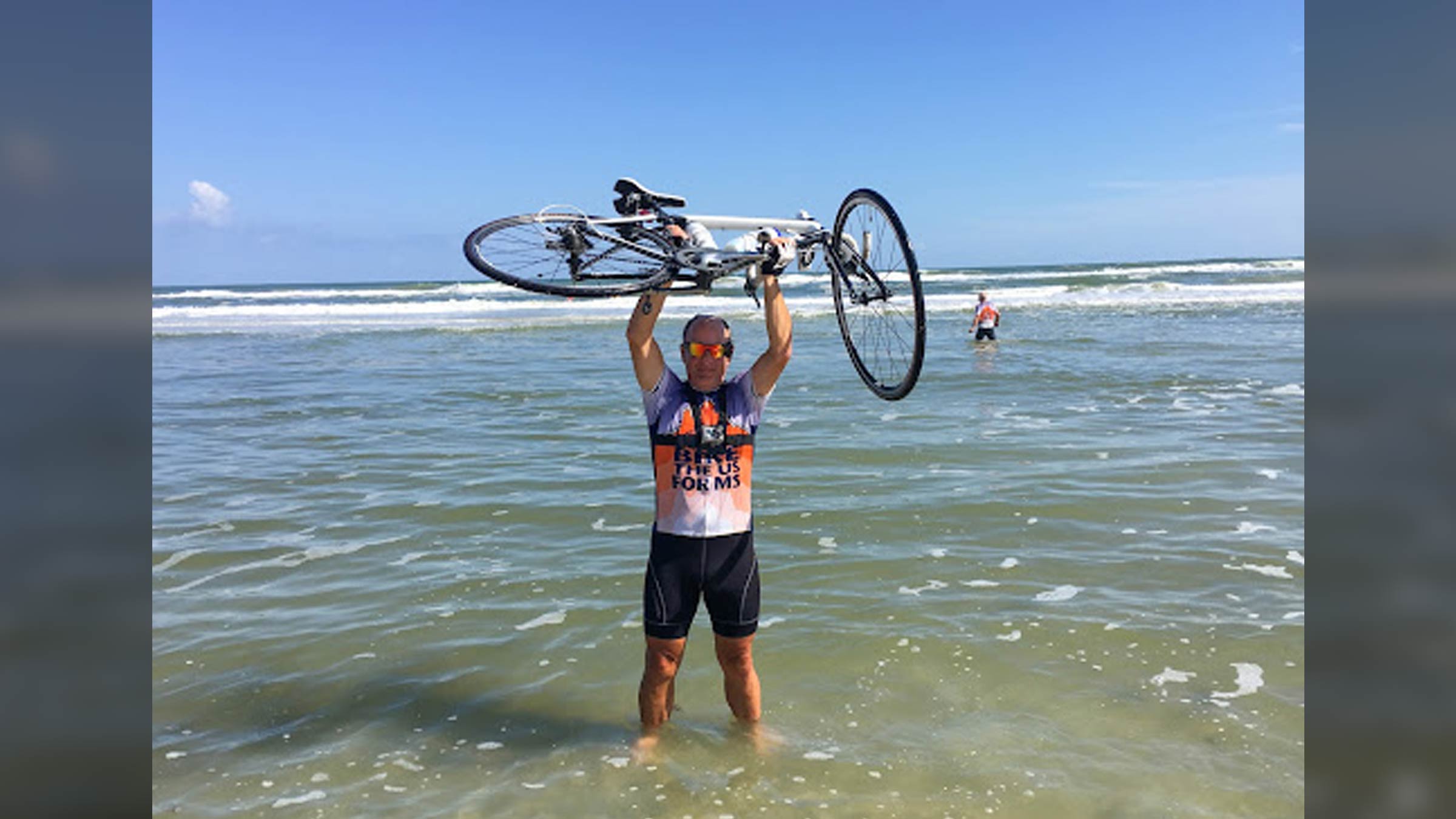 Angel Kowalski, a cancer survivor and avid cyclist, holds his bike above his head on the beach