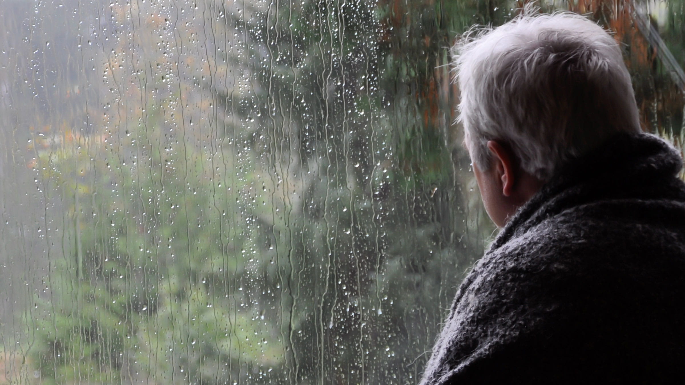 An older man with arthritis looking out the window at the rain