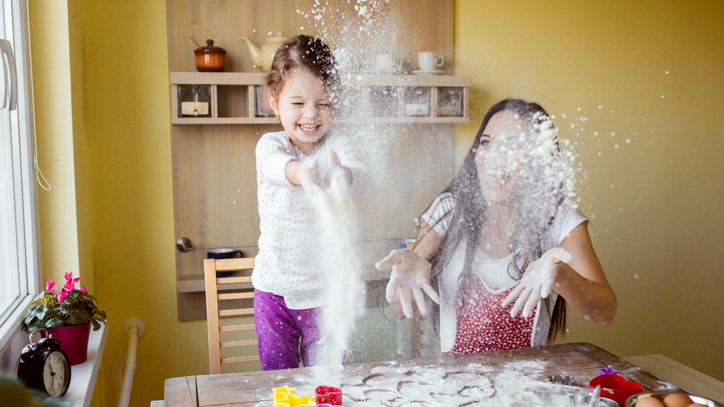 A woman and her young daughter throw flour in the air as they make cookie shapes