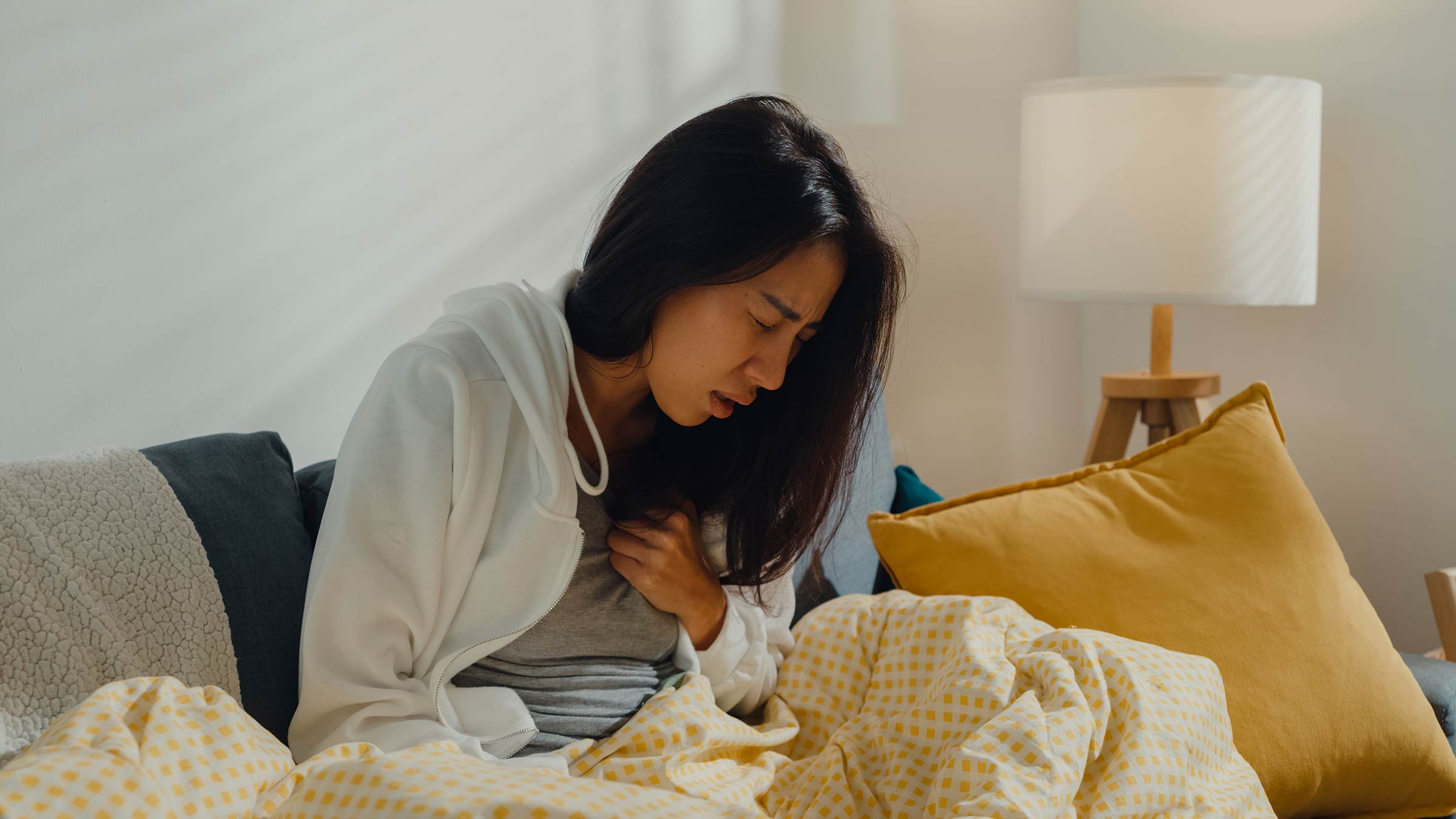 Young woman struggles to cough while sitting on the sofa under a blanket