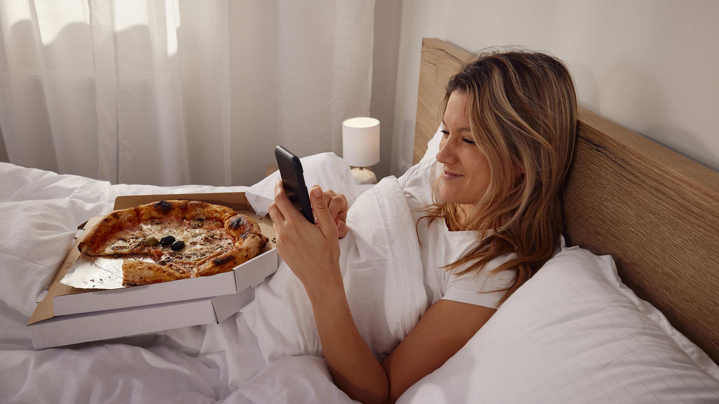 A young woman relaxes in her bed by scrolling on her phone and eating pizza