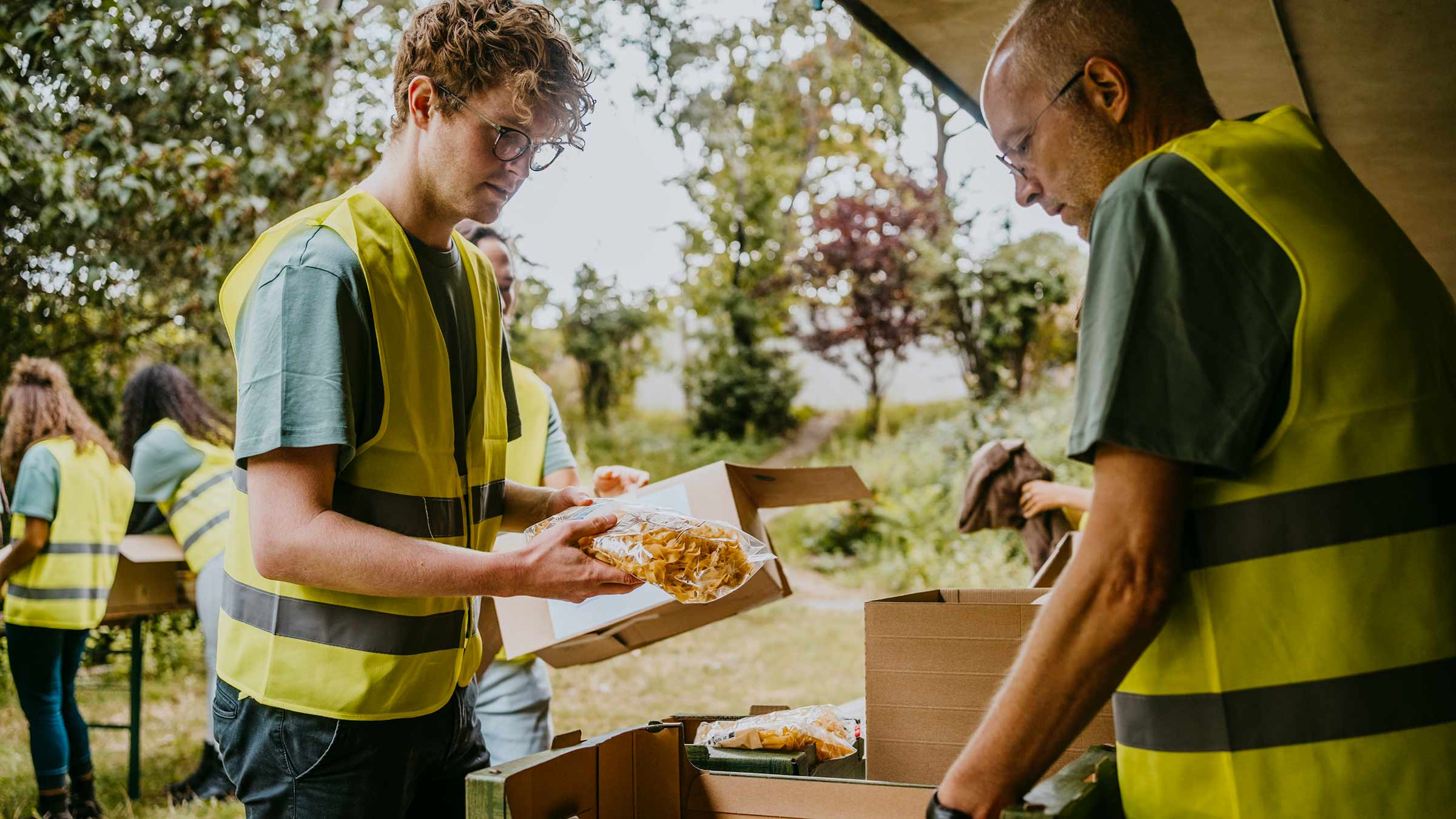 Two men wearing yellow vests pack food packets into a box