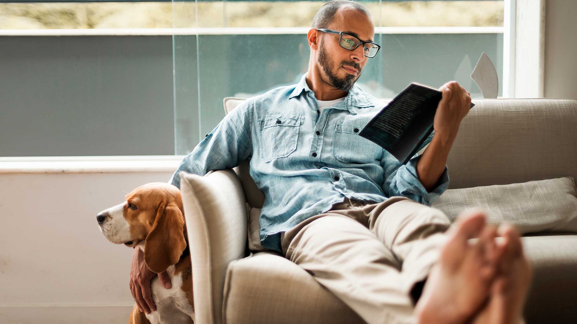 Man sitting on a couch reading and petting his dog beside him