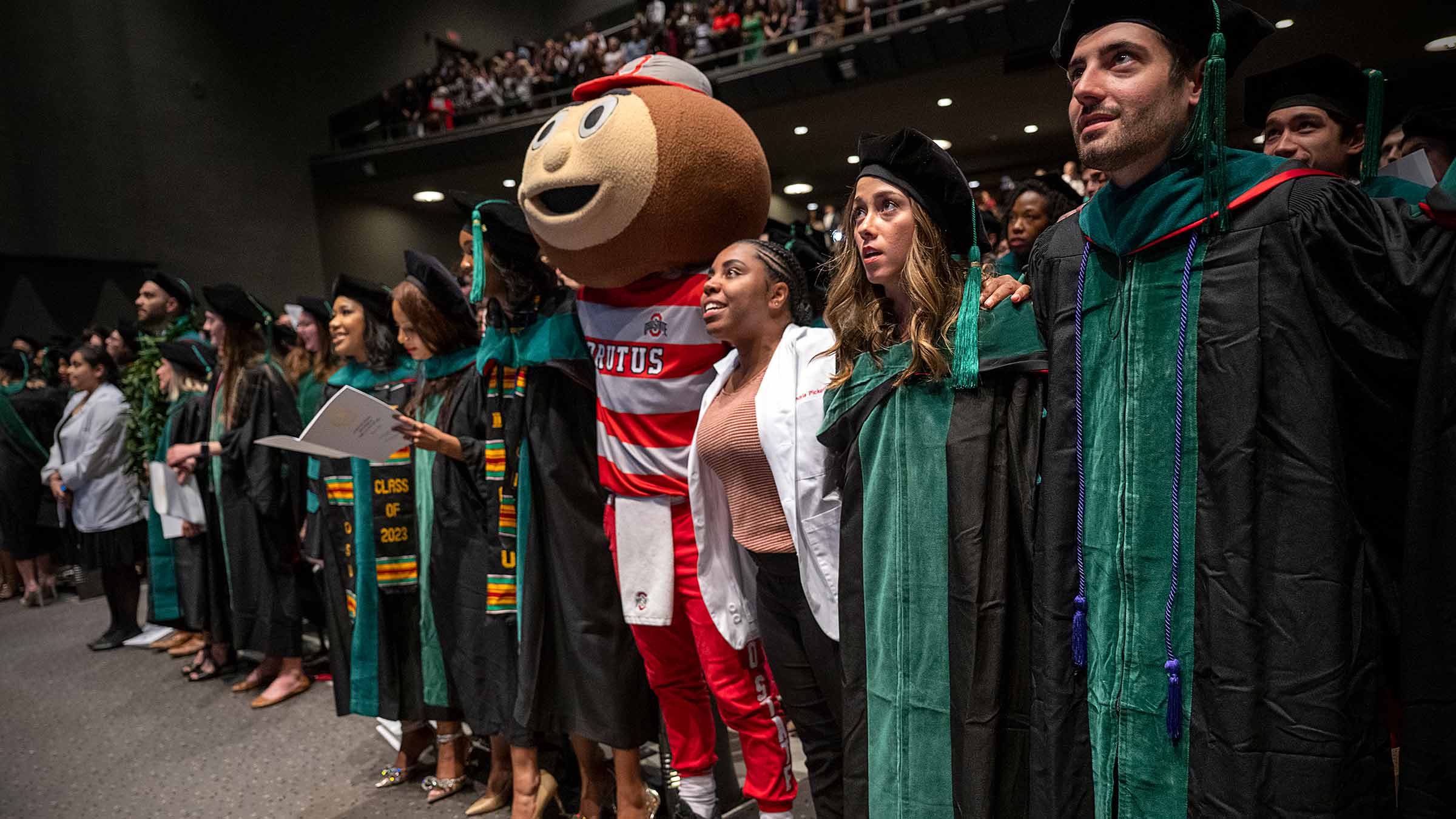 Medical students with interlocked arms sing the Ohio State alma mater at the Doctoral Convocation ceremony with Brutus
