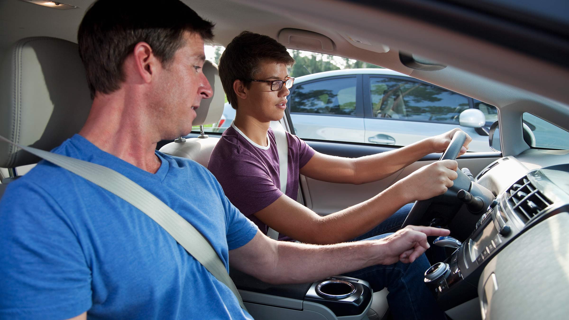 A teen wearing glasses grips a steering wheel sitting in a car’s driver’s seat, while an instructor points from the passenger seat
