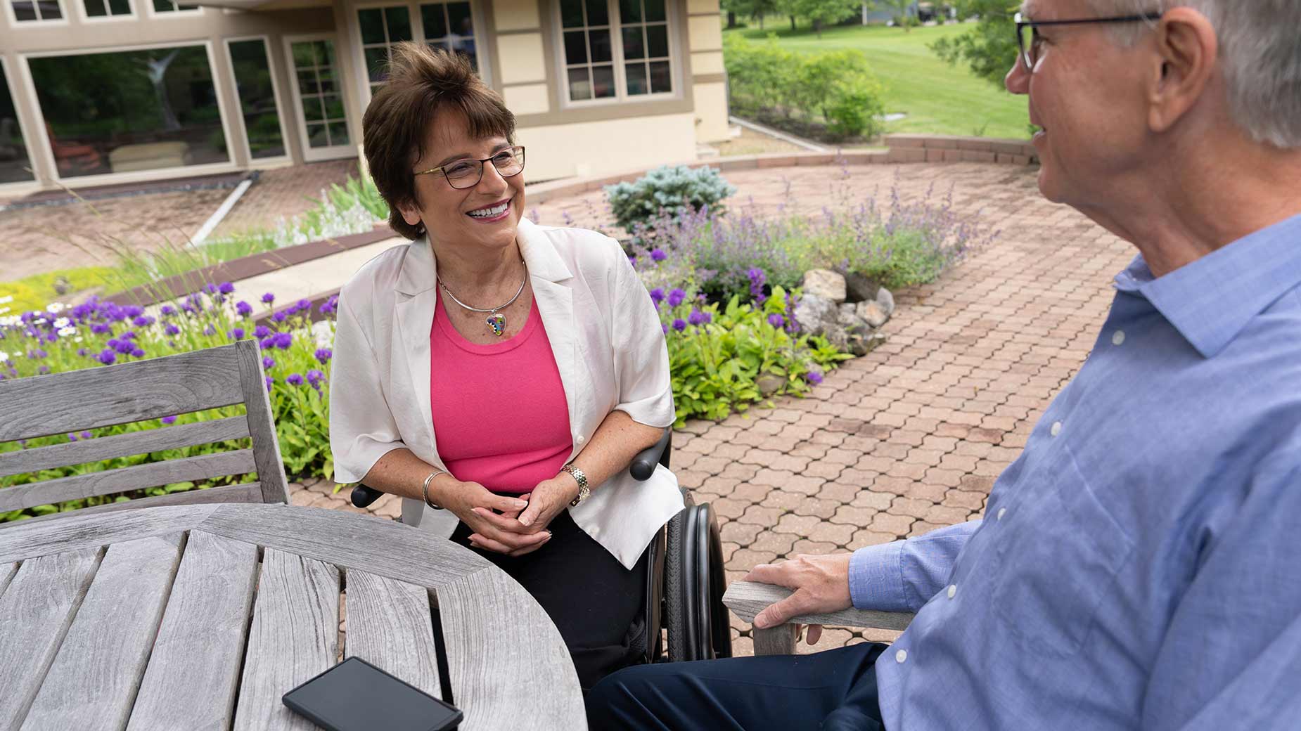 Rosemarie Rossetti sits in her wheelchair at a table outside talking with her husband. 