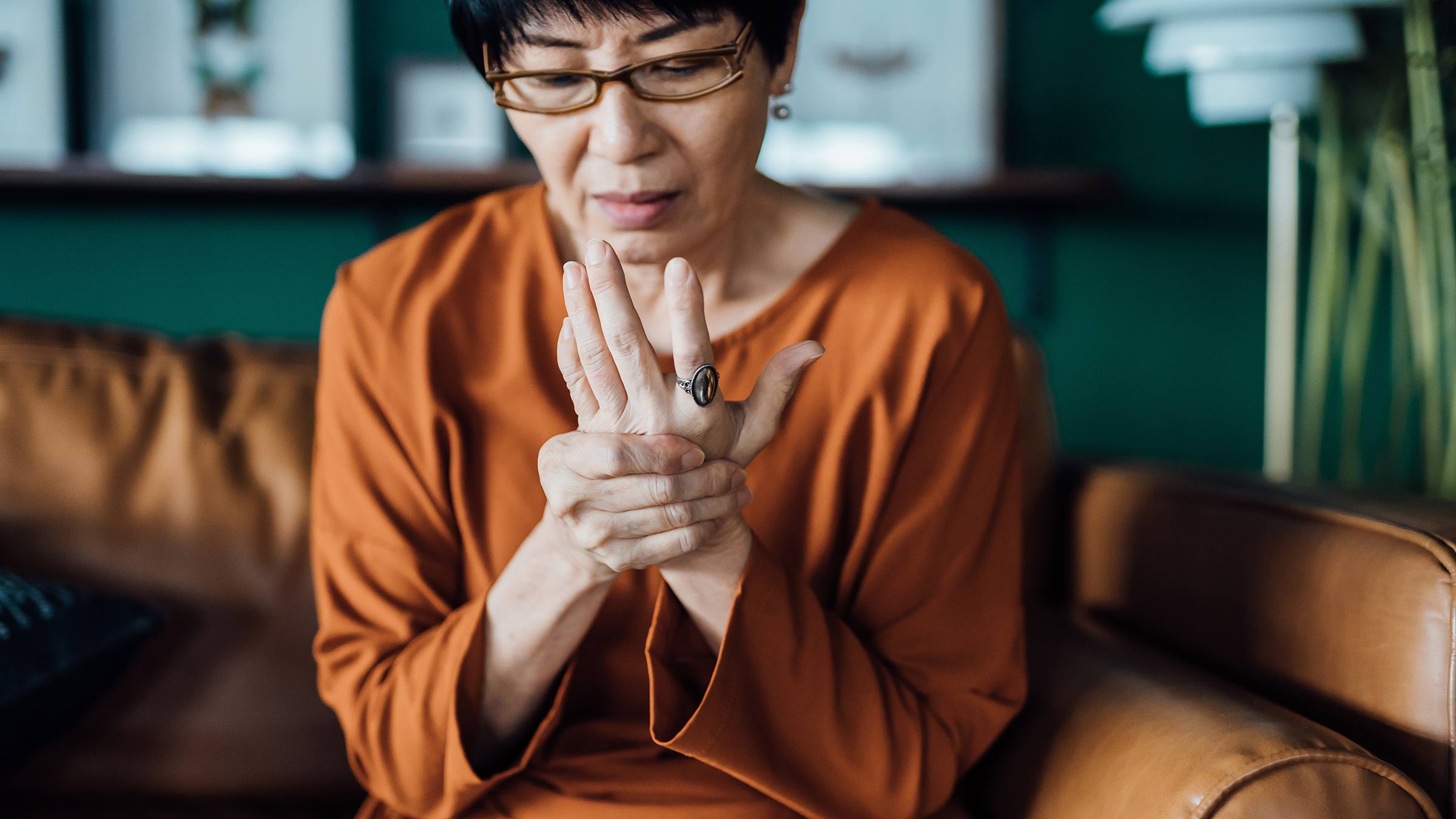 A senior Asian woman sits on a couch while rubbing her hands in discomfort