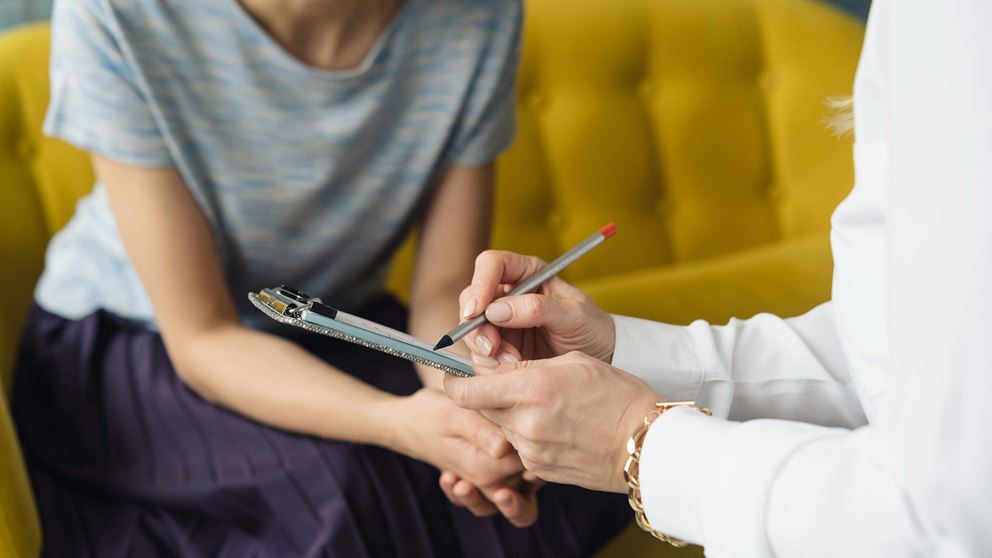 A physician writes on a tablet while talking with a patient who sits on a couch