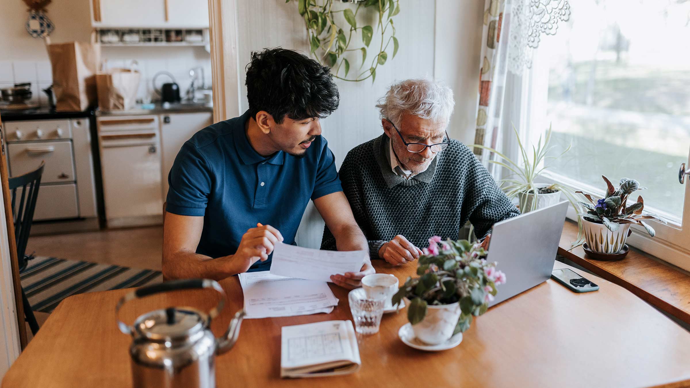 A father and an adult son sitting at a table looking at the documents and a laptop