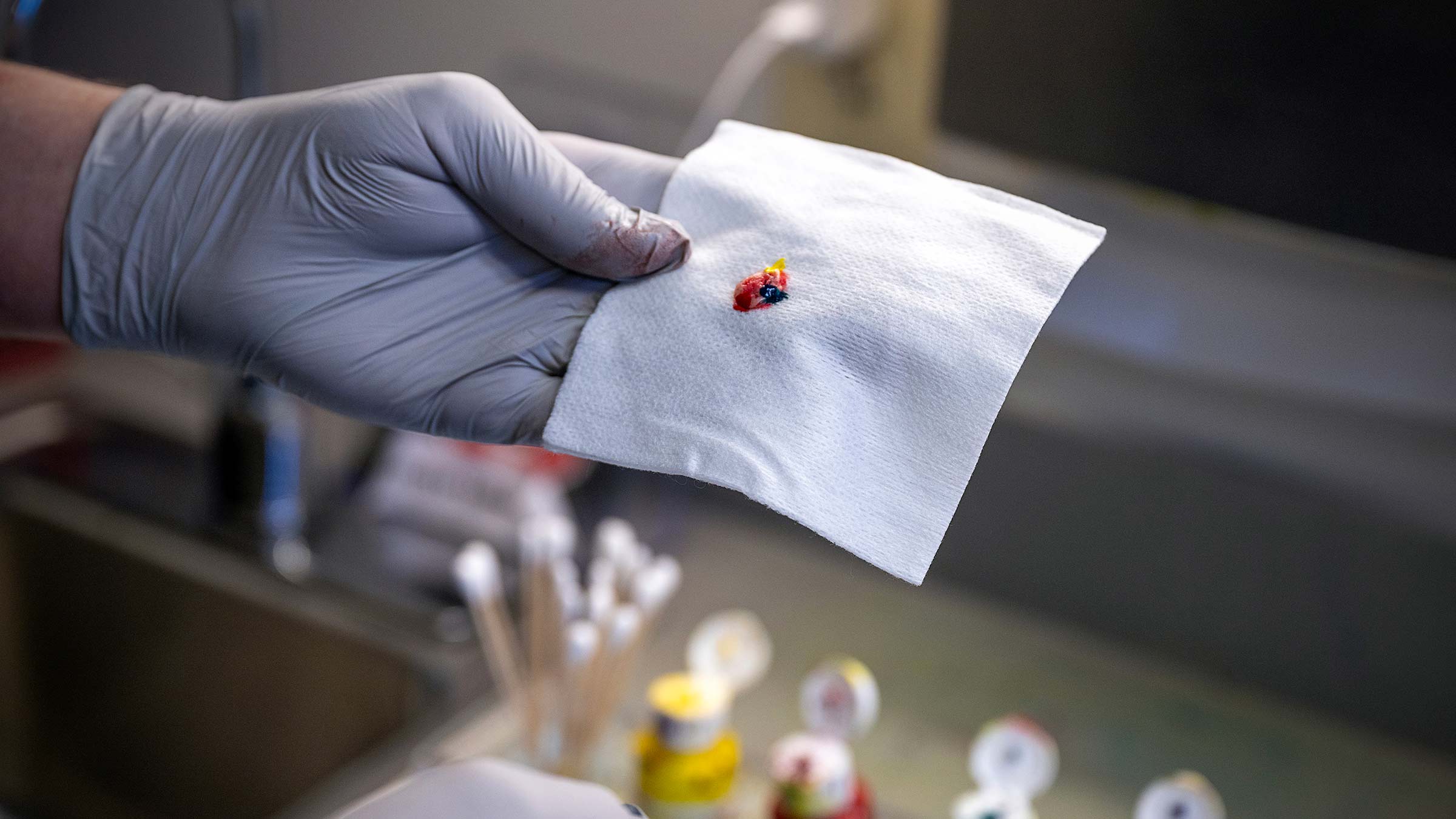 A gloved hand holding a piece of gauze with a multicolored sample on it