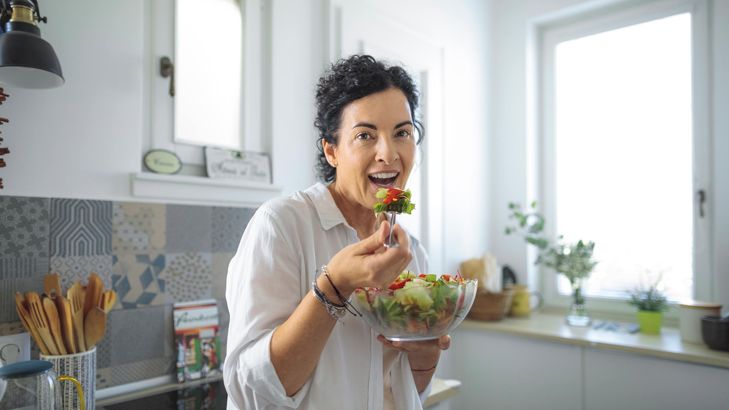 A woman standing in her kitchen and eating a salad