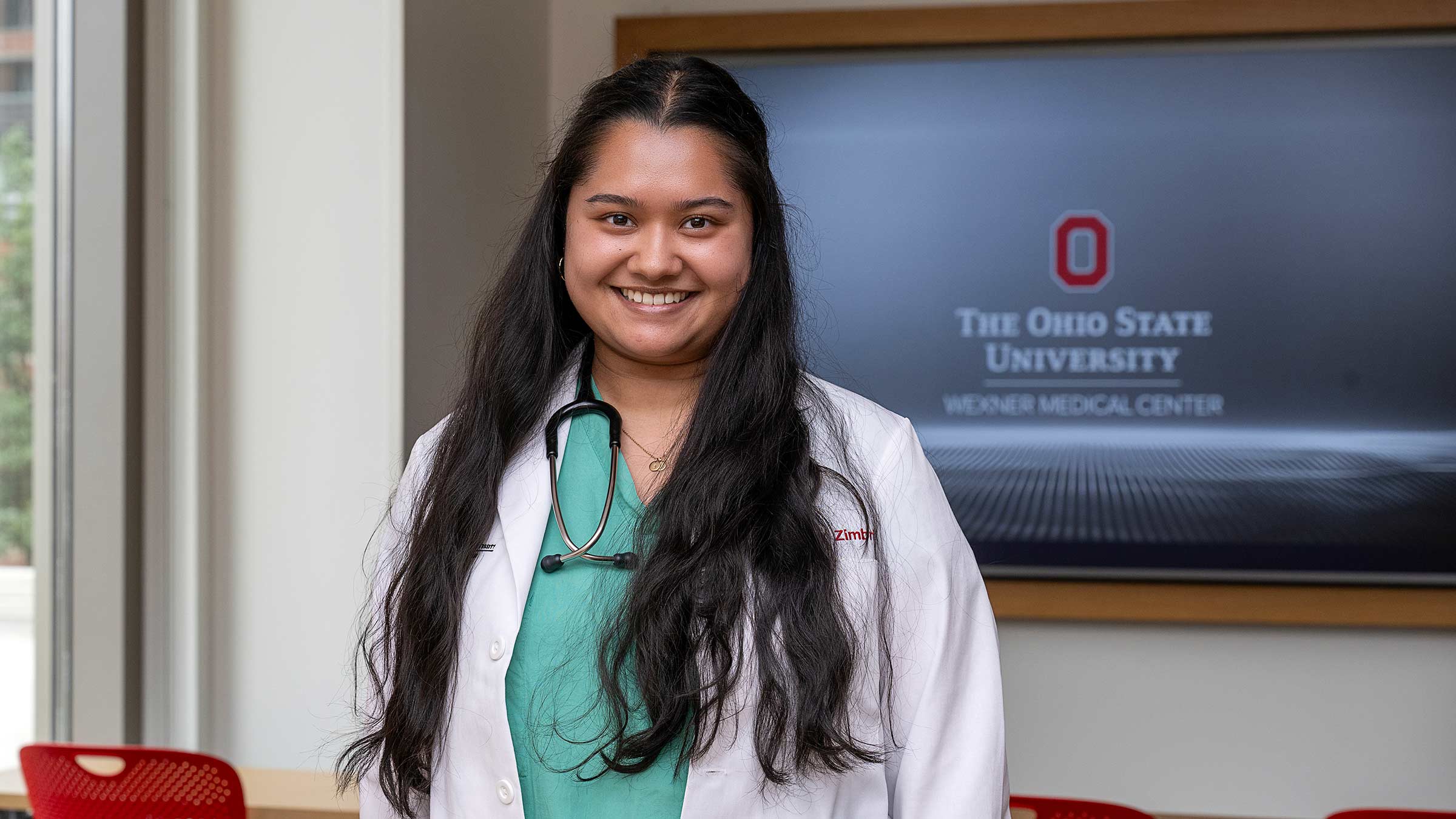Medical student, Jane Chakraborty, standing in a classroom in her medical uniform with a stethoscope around her neck
