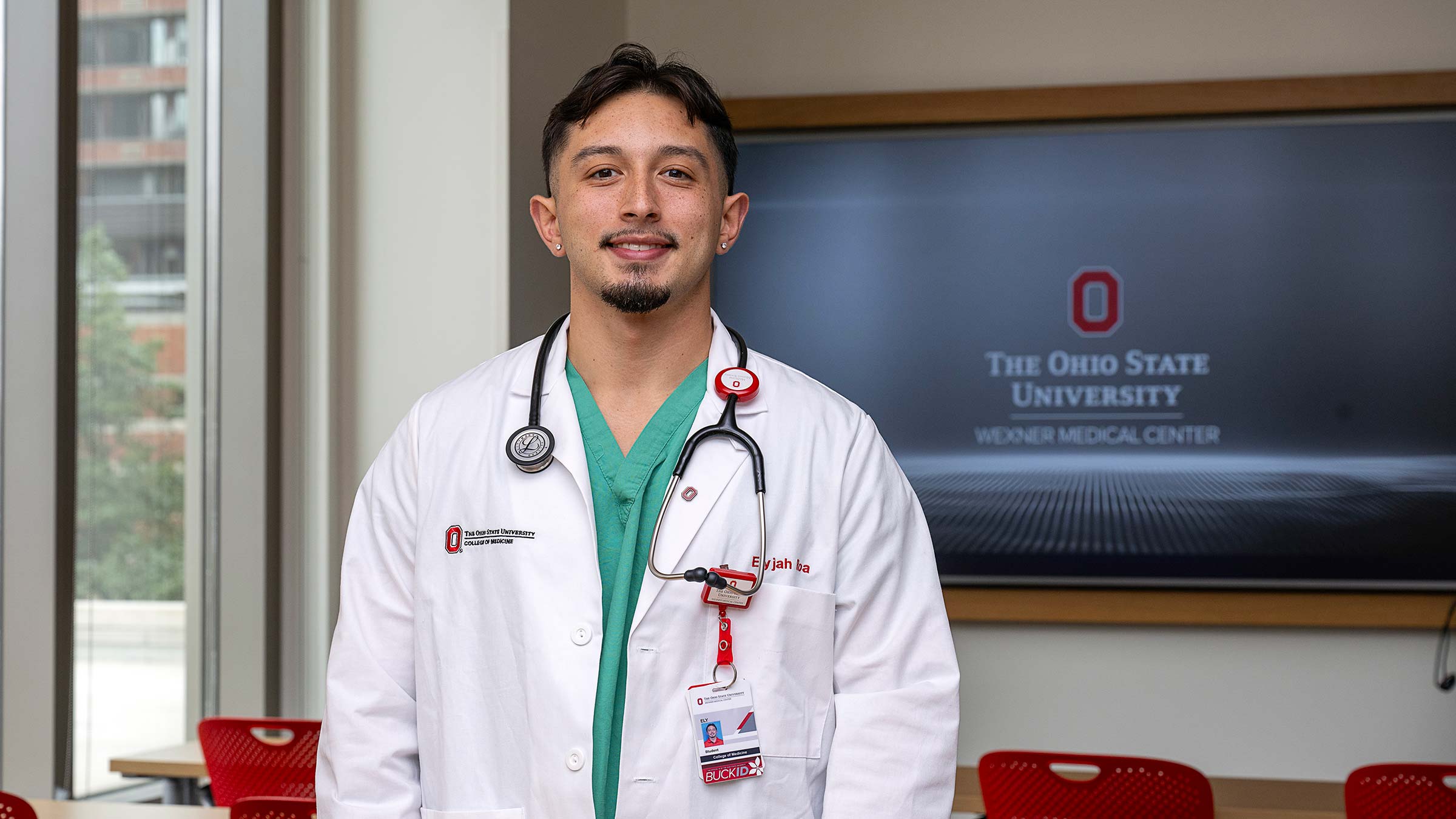 Medical student, Ely Roa, standing in a classroom in his medical uniform with a stethoscope around his neck