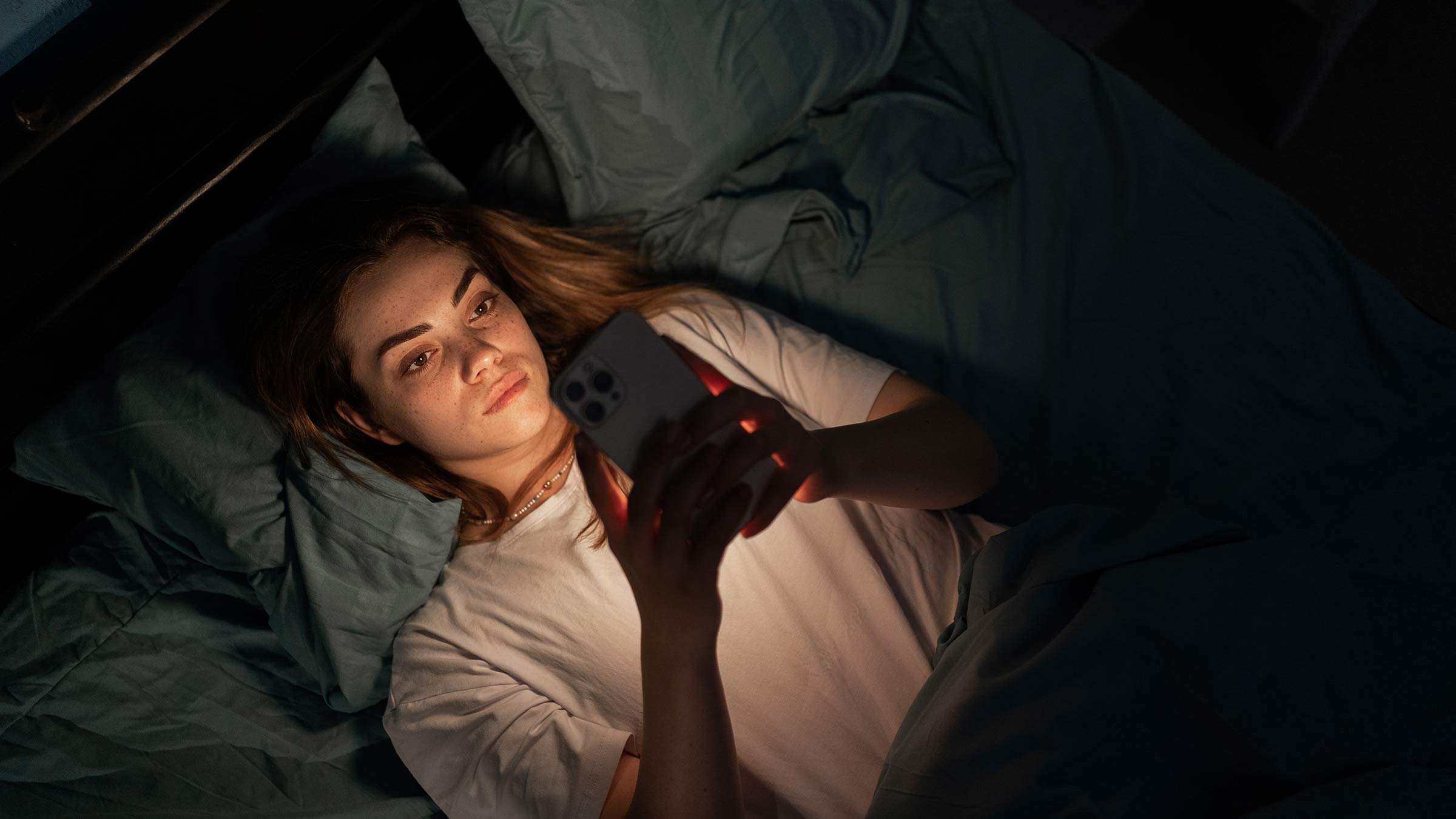 A young woman lying in bed in the dark looking at a smartphone