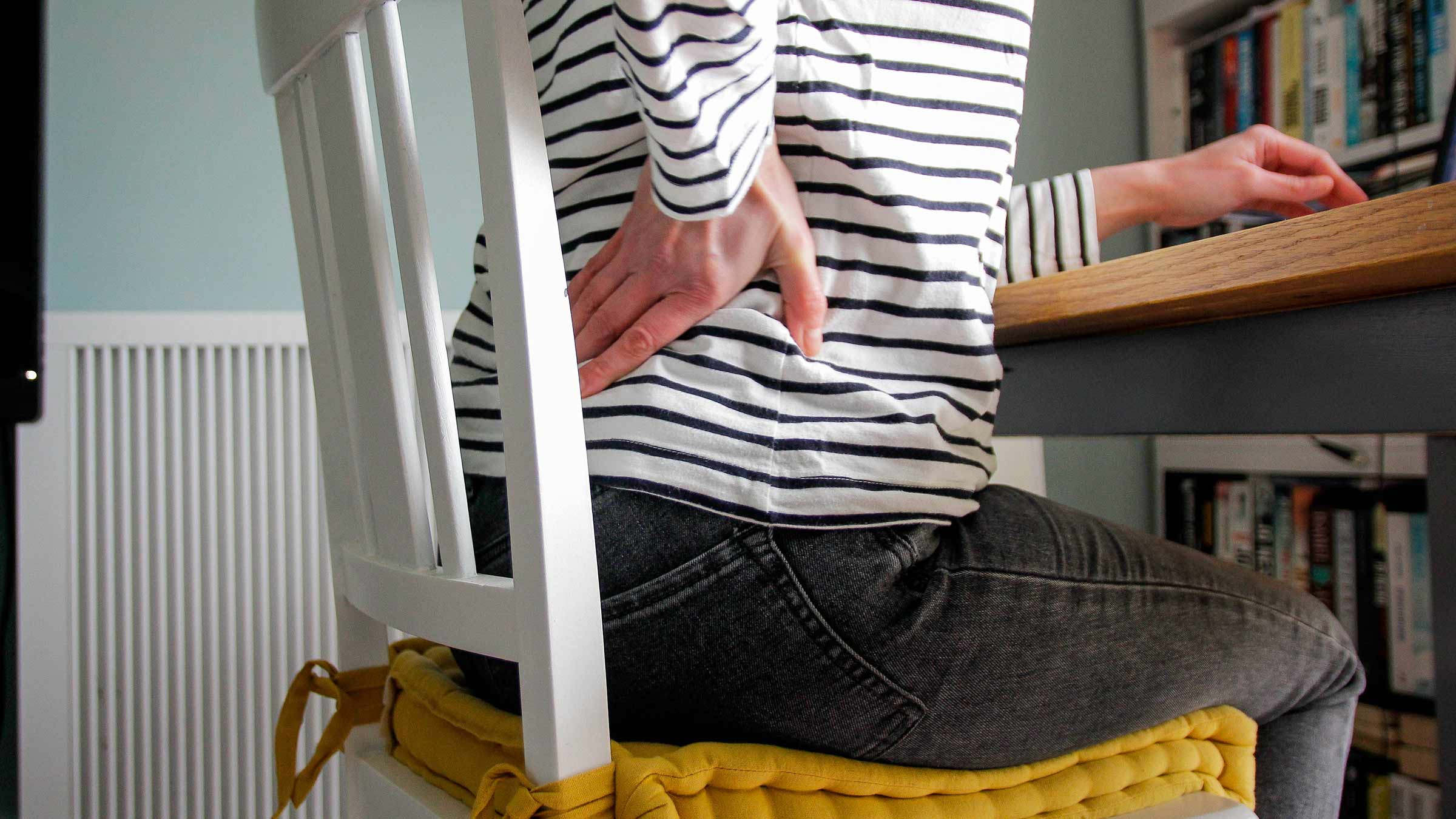 A person sitting at a desk chair with their hand rubbing their back