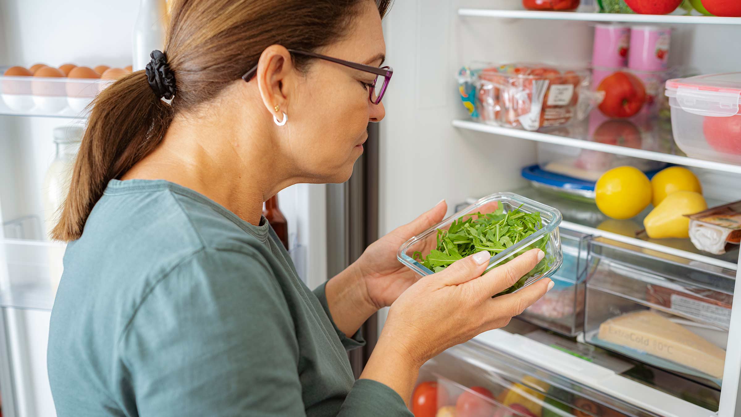 A woman smelling a container of greens from her refrigerator