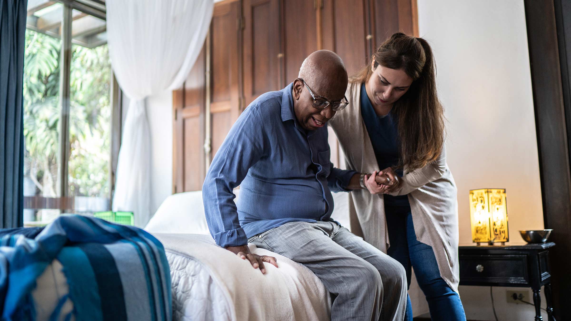 A young woman helping an elderly man get off the bed