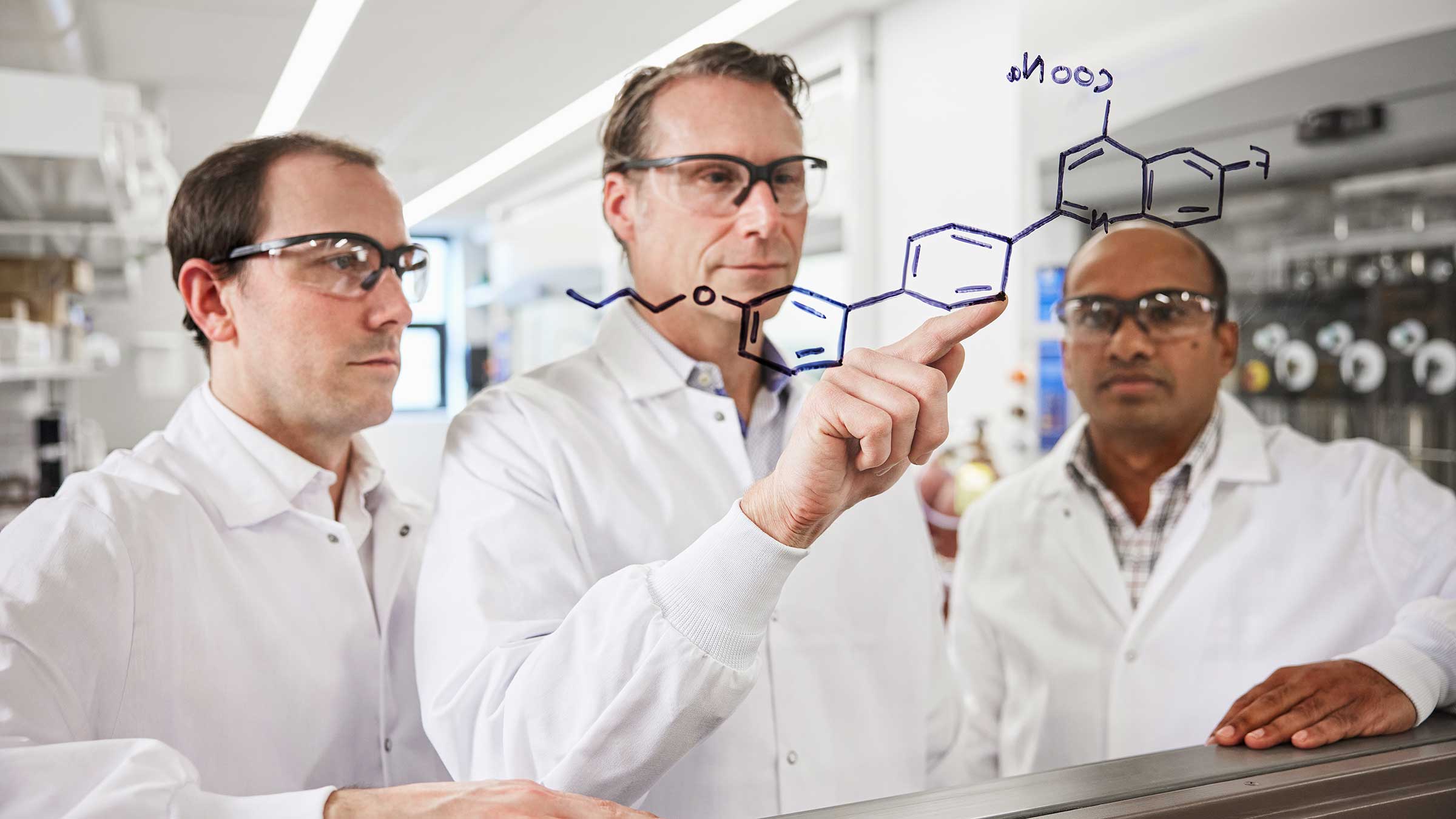 Three scientists in lab coats examining a transparent board with a chemical formula