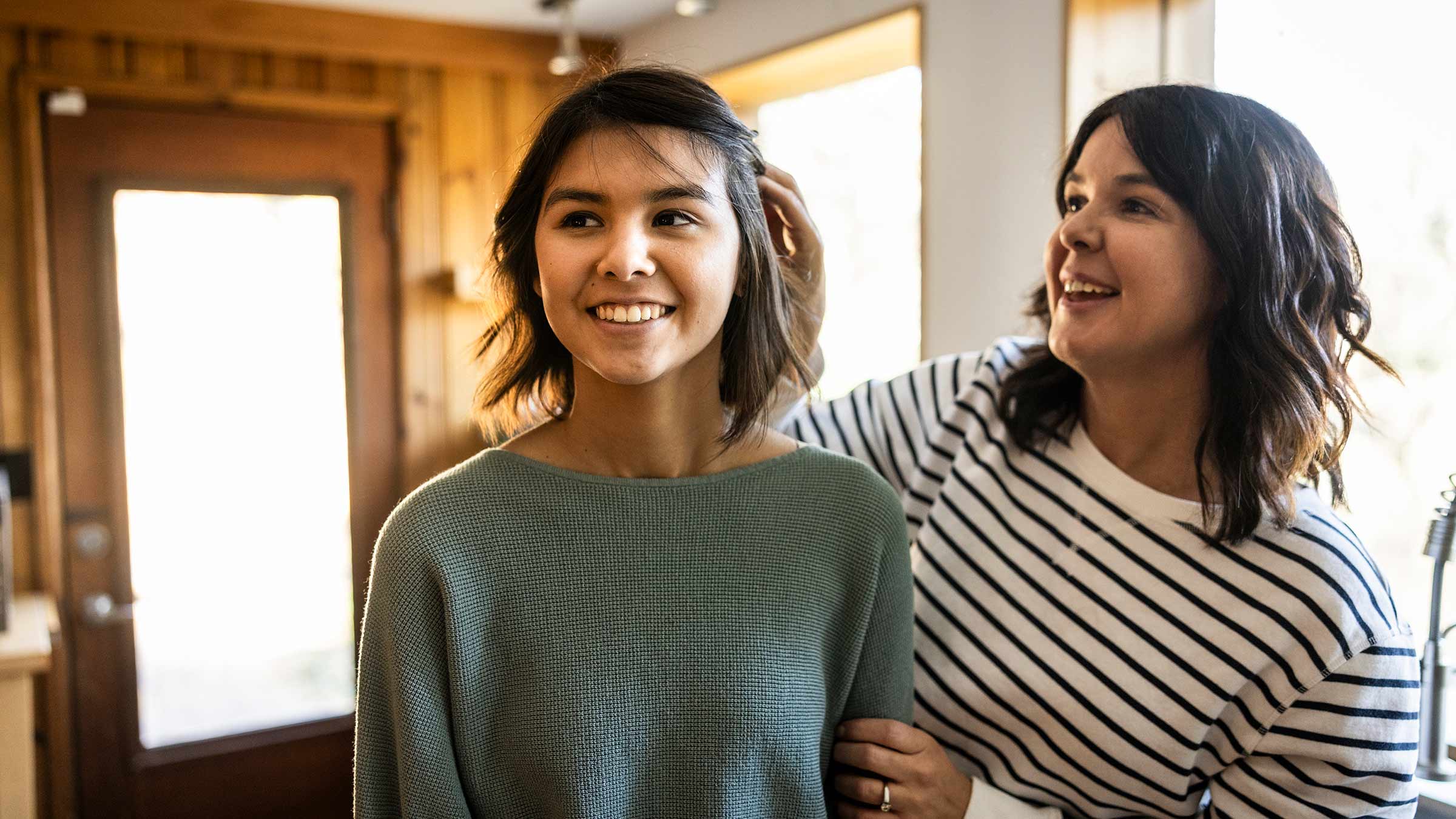 A mother fixing her teen daughter’s hair in a kitchen