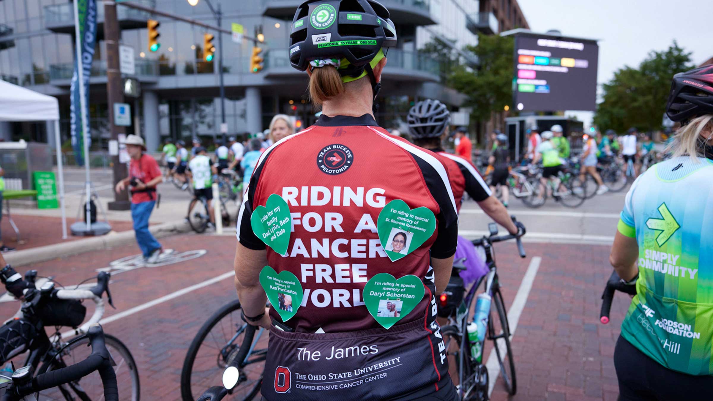 A rider wears a jersey that says 'Riding for a cancer-free world,' paper hearts with names pinned on it