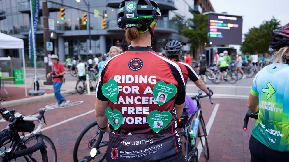 A rider wears a jersey that says 'Riding for a cancer-free world,' paper hearts with names pinned on it
