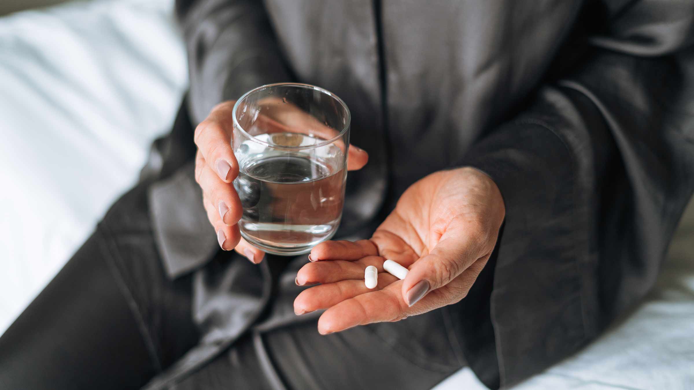 A person sitting on a bed holding a glass of water in one hand and pills in another