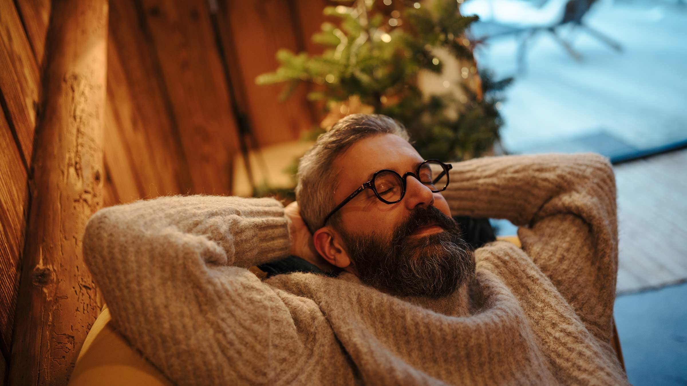 A man relaxing on a sofa with his eyes closed and his hands behind his head, with a holiday tree in the background