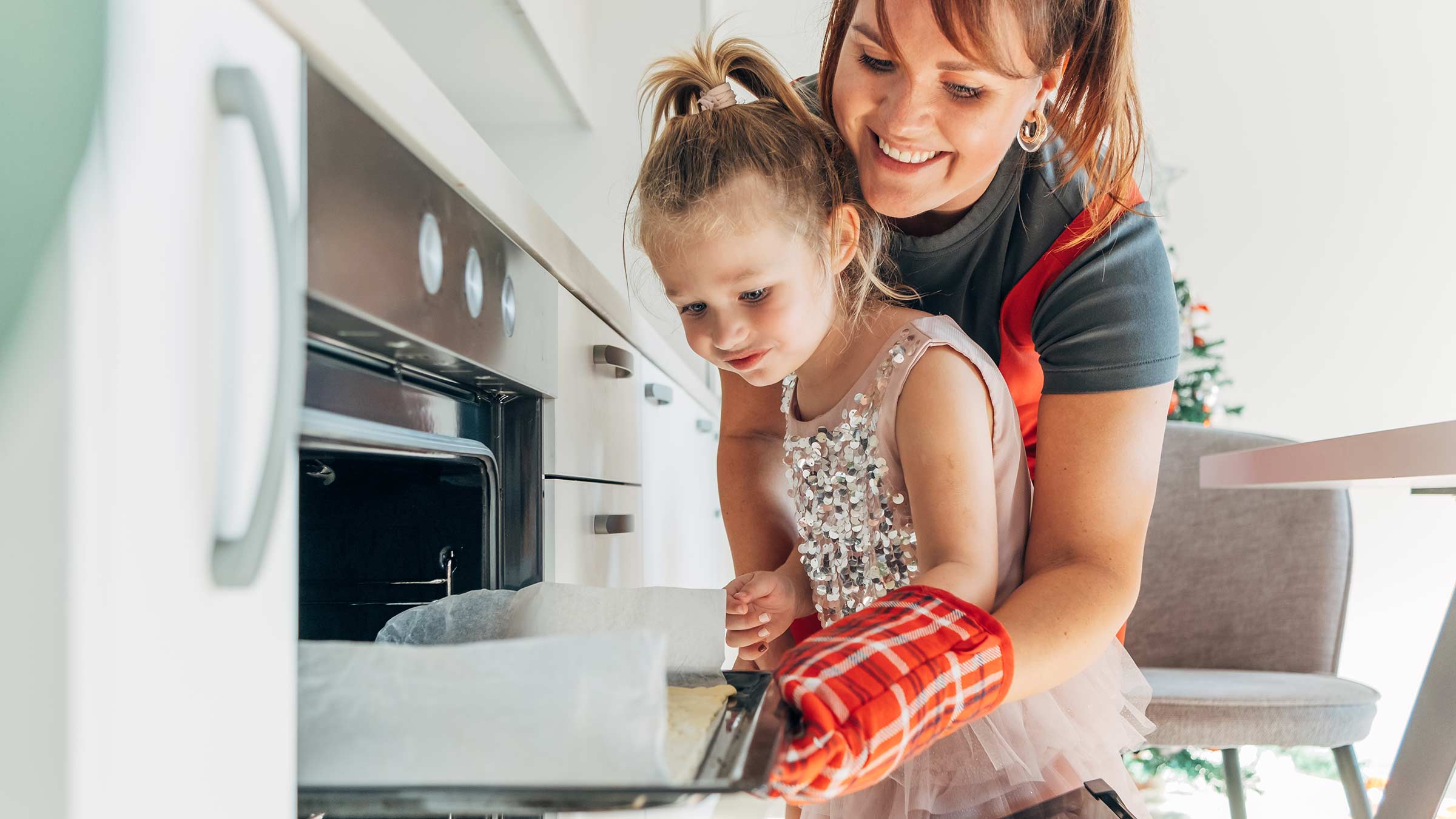 A mother helping her young daughter with putting baked goods in an oven