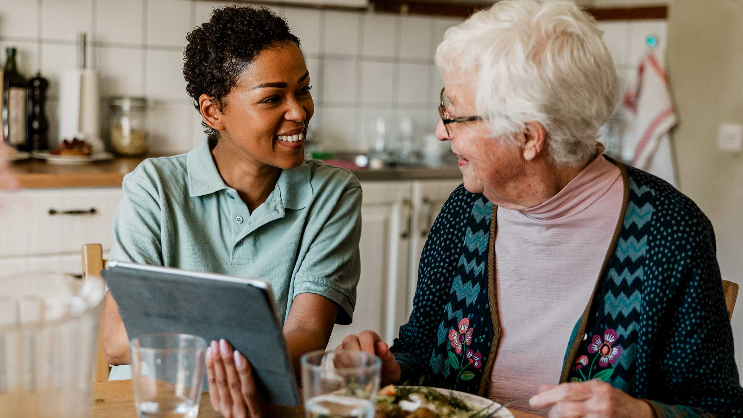 A caretake helps a senior woman use her tablet at the breakfast table
