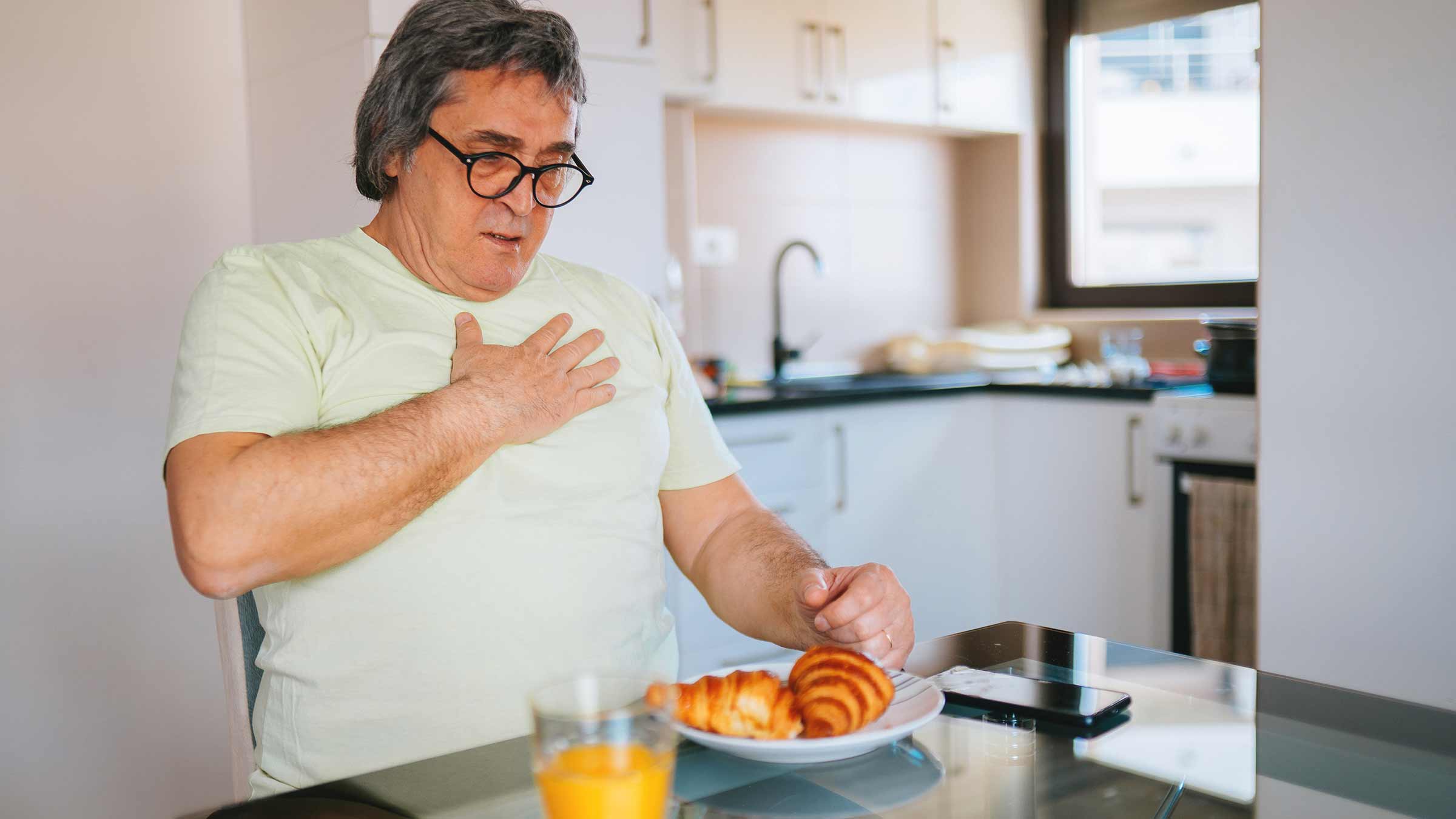 A person sitting at a kitchen table with food, clutching their chest in pain