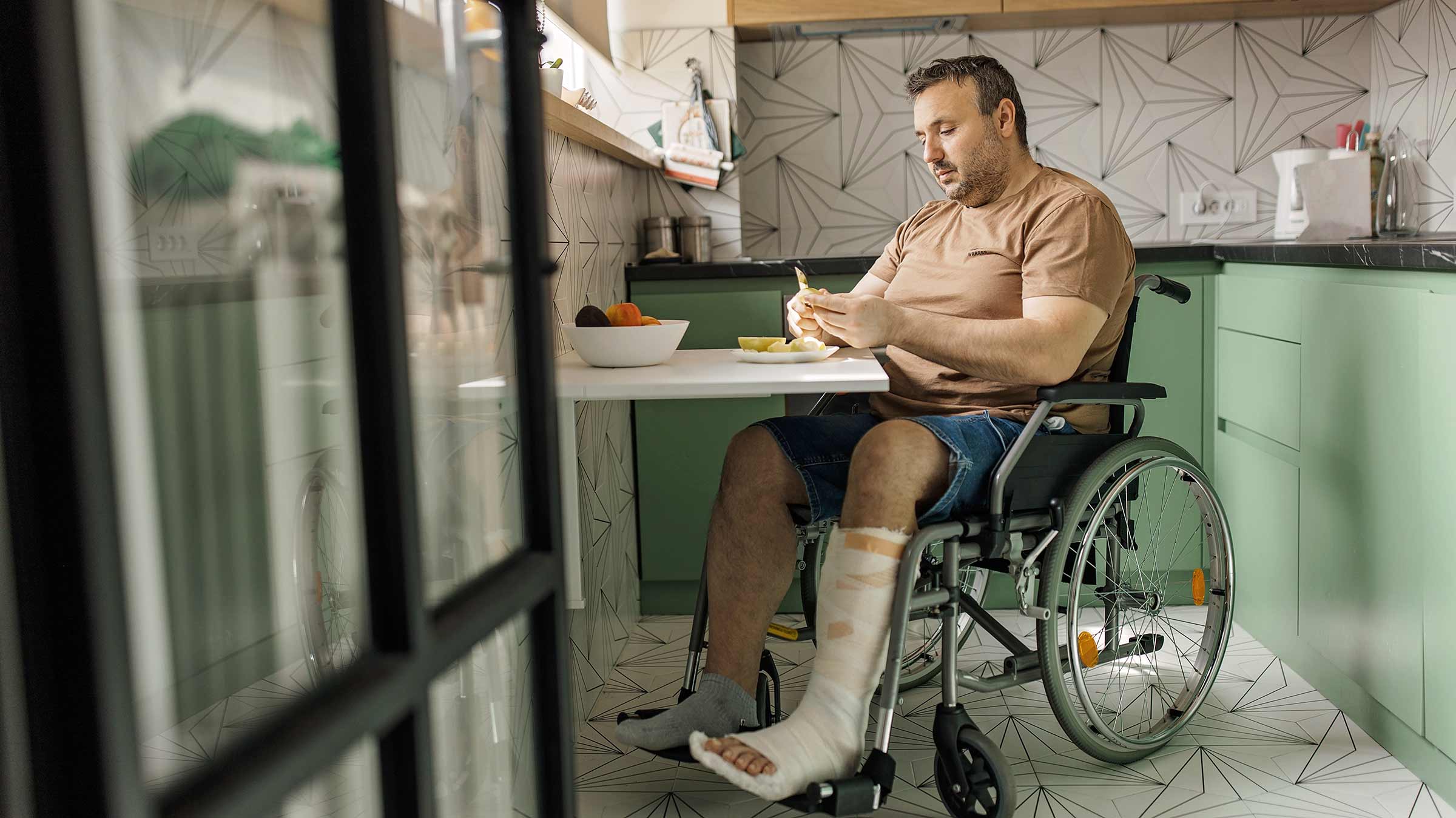A man sitting in a wheelchair with a leg cast eats breakfast at a kitchen table