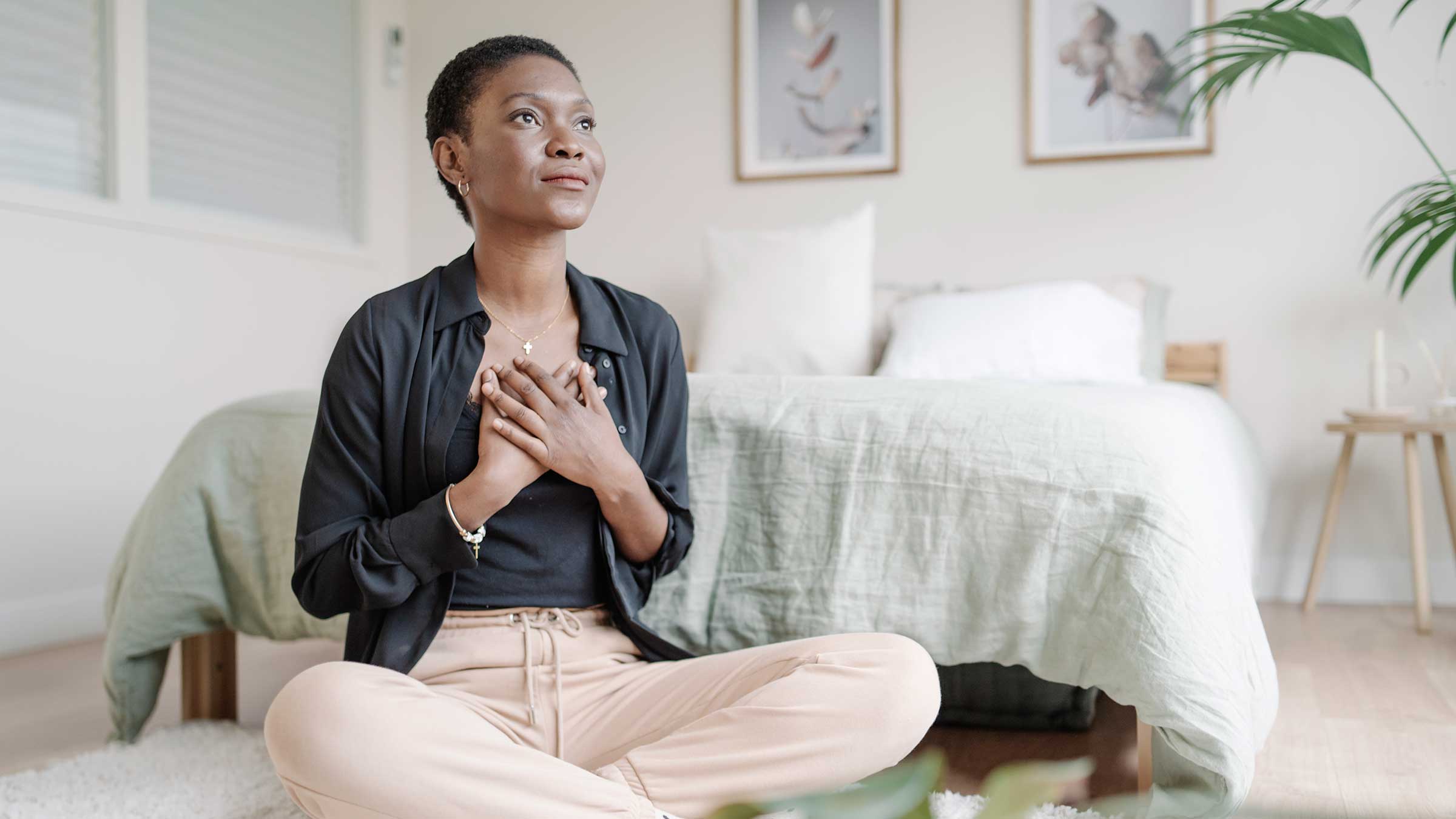 A Black woman sitting on the floor in her bedroom with her hands held across her chest