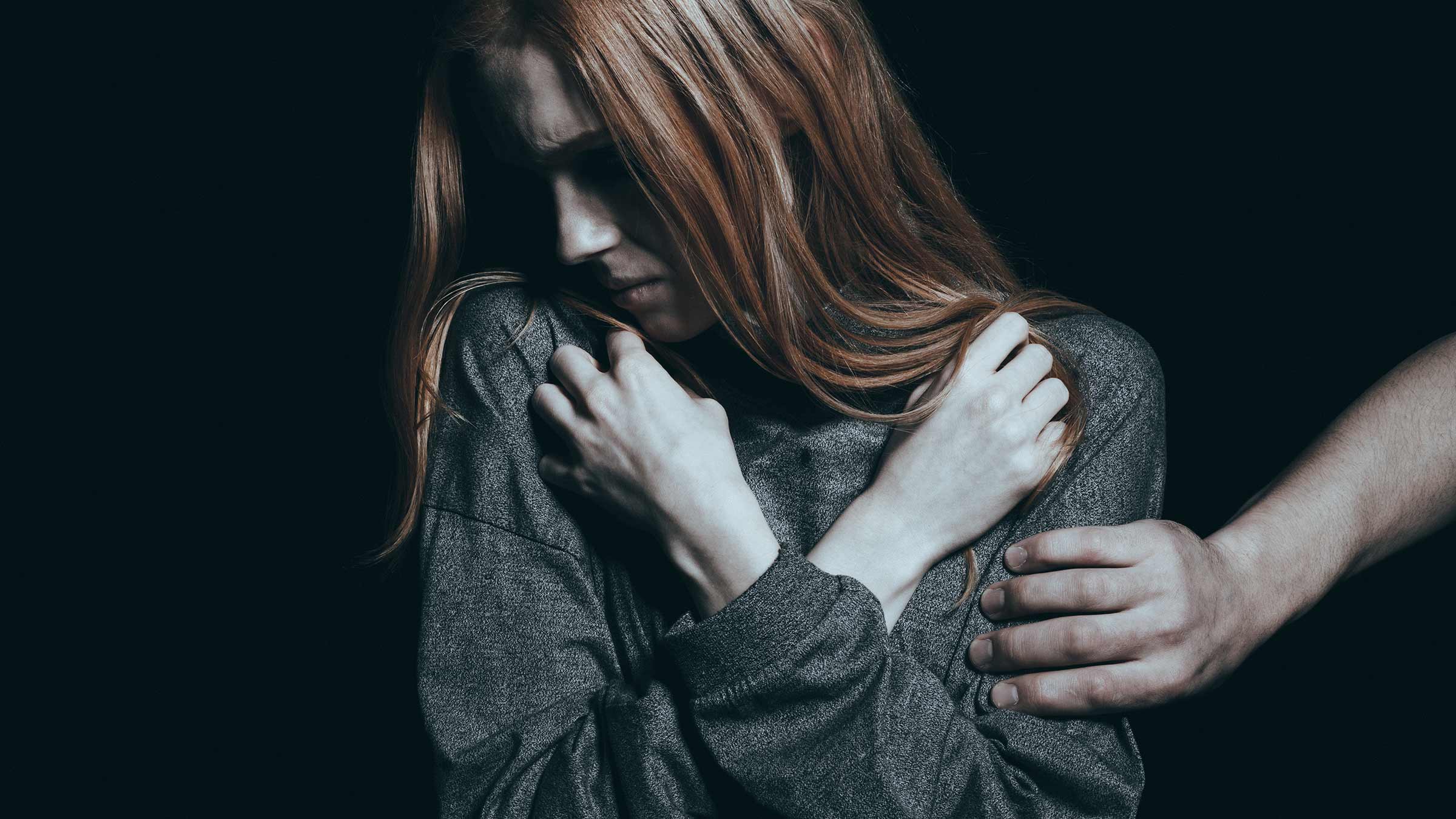 A young woman hiding her face and showing discomfort with a stranger touching her, with an all-black background